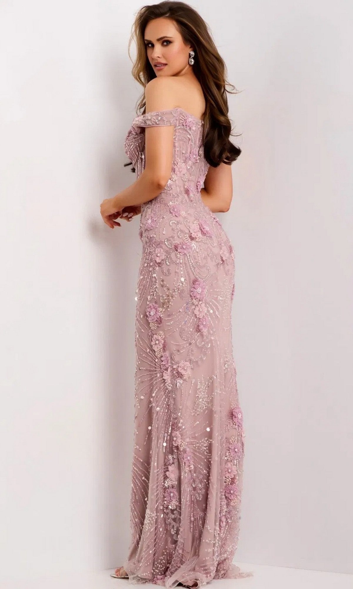 A woman poses against a plain light background, looking back over her shoulder while wearing the Jovani 44007 long prom dress—an off-the-shoulder, fitted pink gown featuring elegant 3D floral appliques.